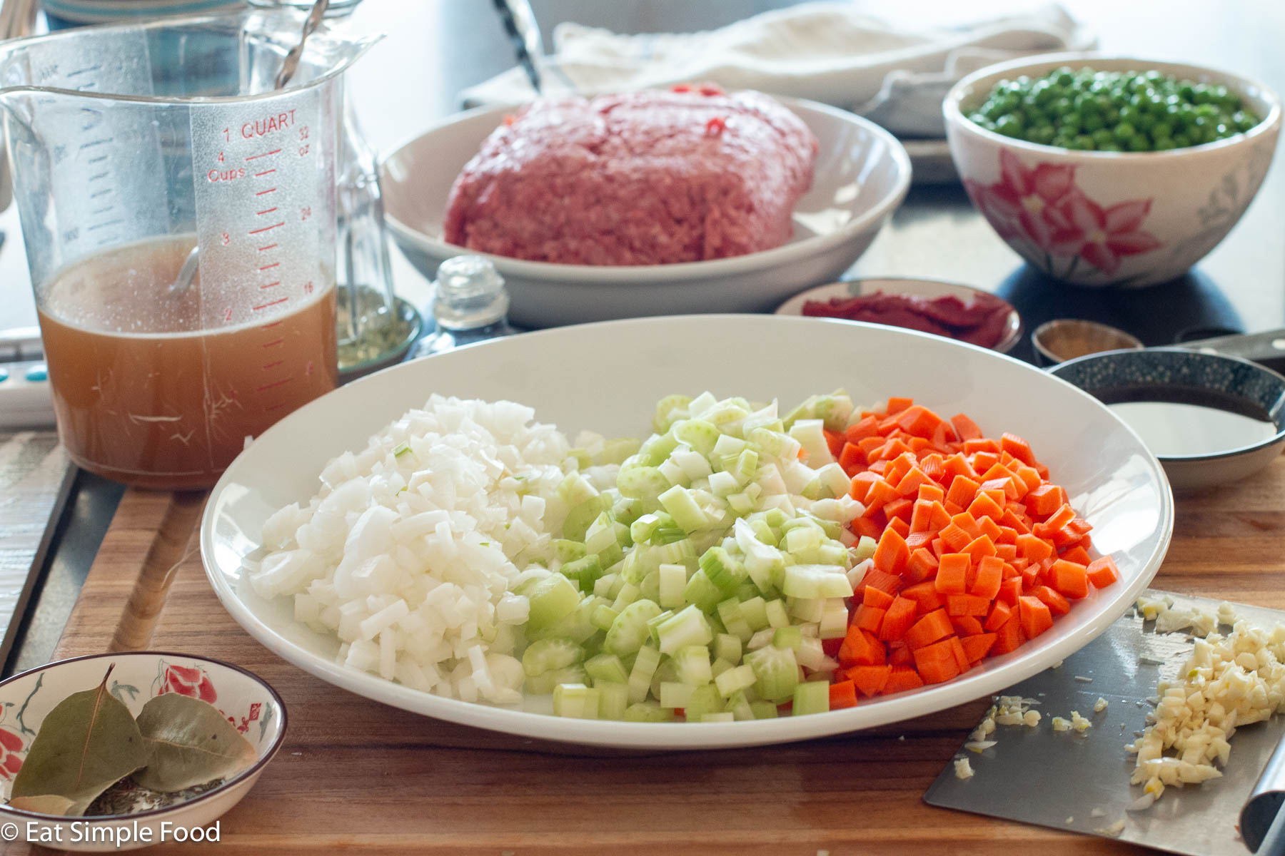 Large white plate with diced onions, celery, and carrots. Plate of ground beef and bowl of raw green peas in the background.