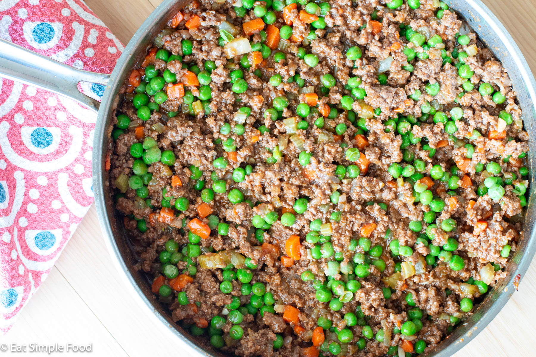 Top down view of a stainless steel pan filled with cooked ground beef and diced cooked carrots and green peas.
