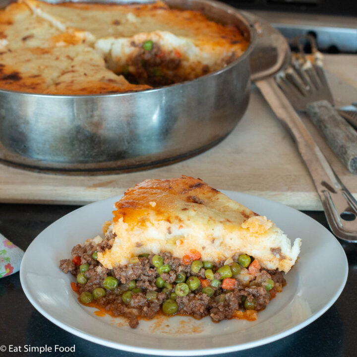 White plate with a piece of meat pie topped with mashed potatoes and a stainless steel dish of a whole casserole in the background. Close up.