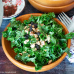 Close up of wood bowl of green salad with raisins, crushed almonds, and red pepper flakes. Fork on the side.