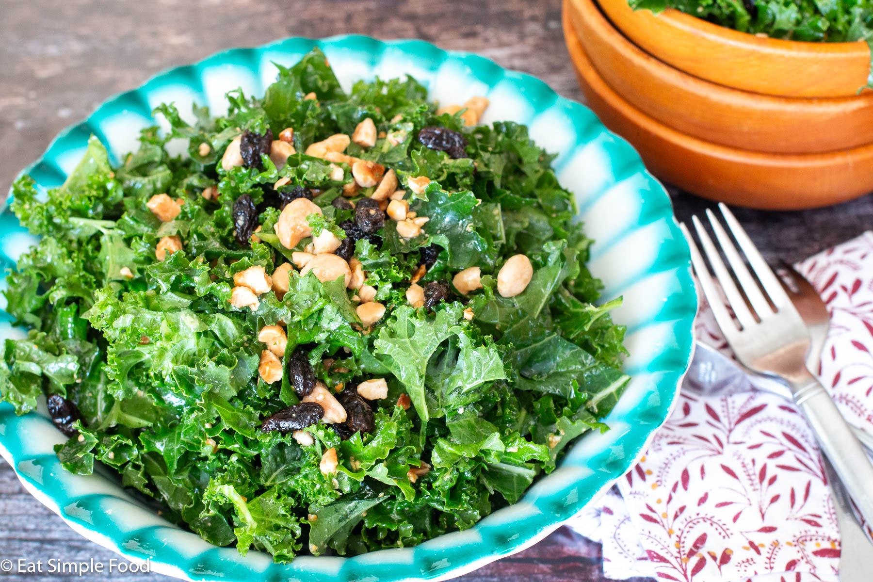Side view of curly kale salad in a turquoise bowl topped with almonds and raisins. Fork and napkin on the side.