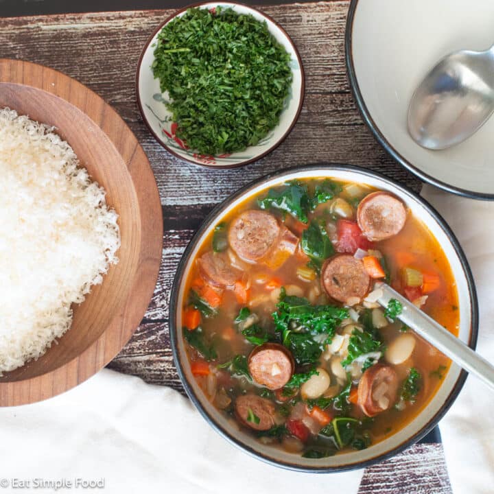 Top down view of white soup bowl filled with broth, kale, beans, and sliced sausage. Spoon sticking out. Grated Parmesan cheese on the side in a wood bowl. Chopped parsley in a small bowl on the side.