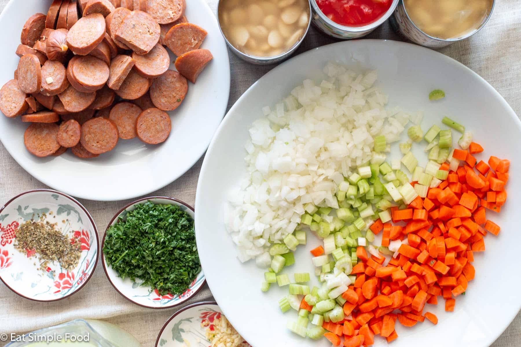 Chopped onion, celery, carrot on a white plate. Sliced smoked sausage on a smaller white plate. Three small bowls of Italian seasoning, chopped parsley, and chopped garlic on the side.
