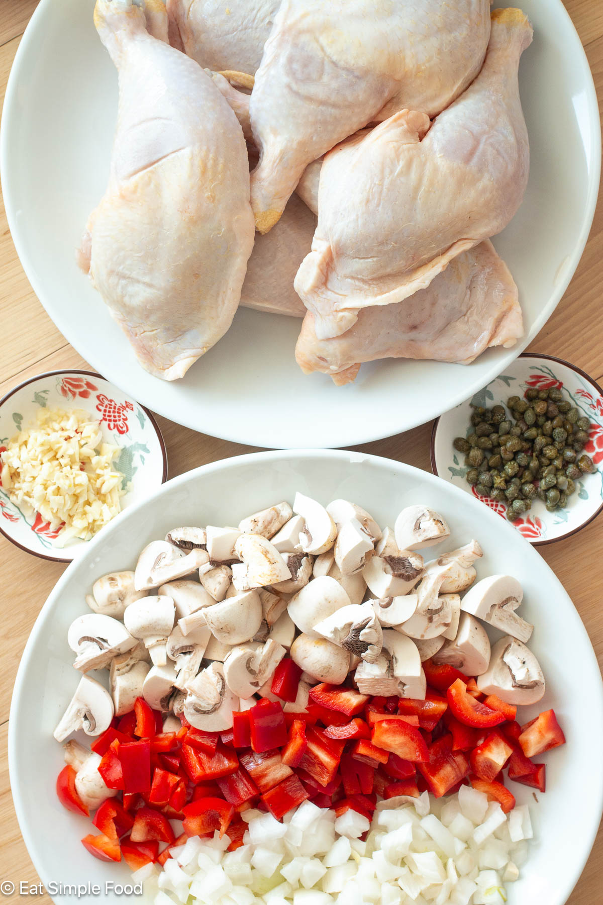 Top down view of ingredients: 5 chicken legs on a white plate; diced mushrooms, red peppers, and onions on a white plate, a small container of capers, and a small container of diced garlic.