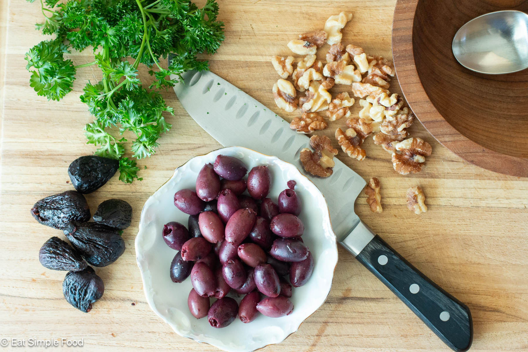 Ingredients on a wood cutting board: kalamata olives bowl, 6 dried figs, handful of walnuts, several sprigs of fresh parsley.