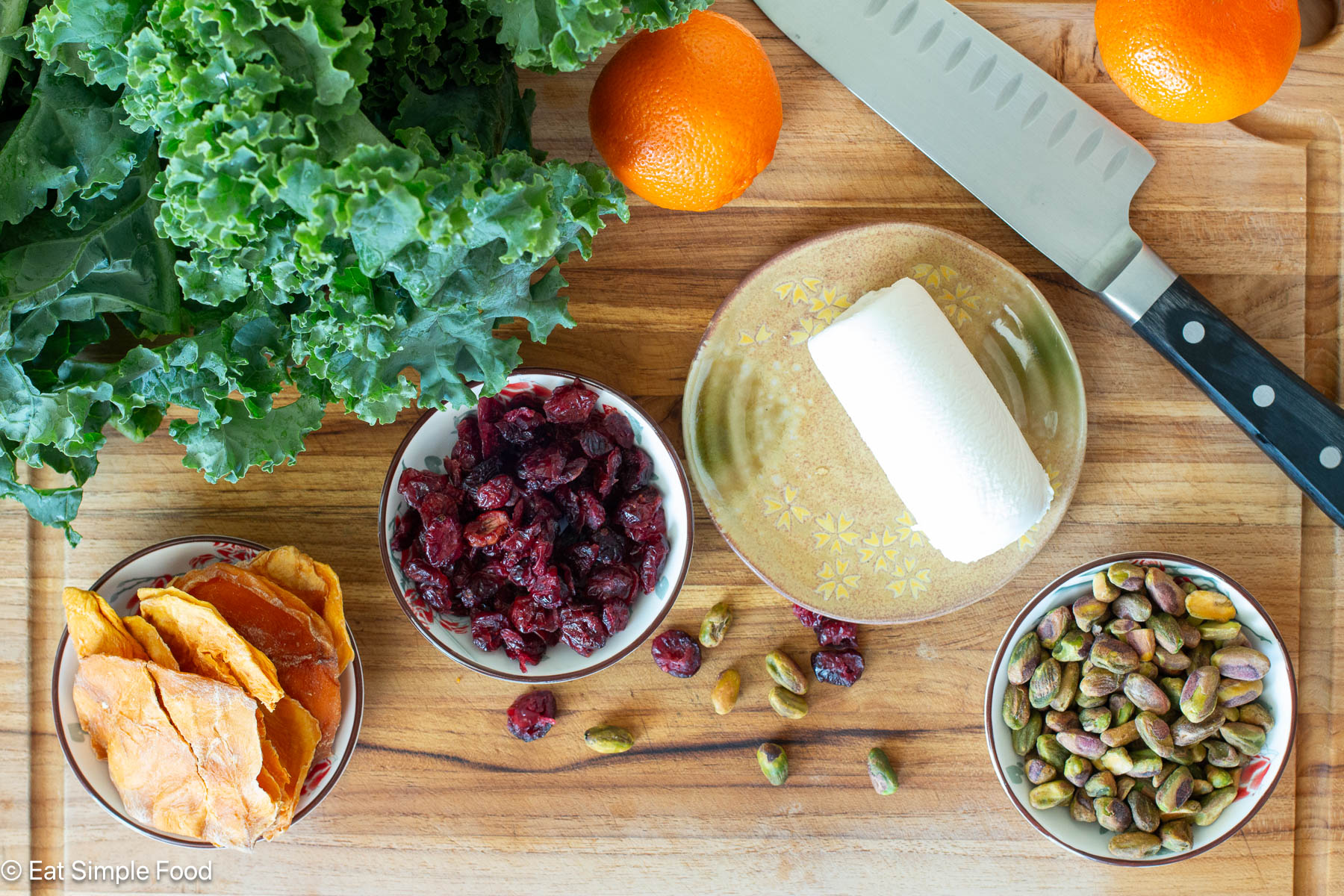 Ingredients on a wood cutting board with a knife. Cranberries. dried mango slices, whole pistachios, a log of goat cheese, 2 oranges, and a head of kale.