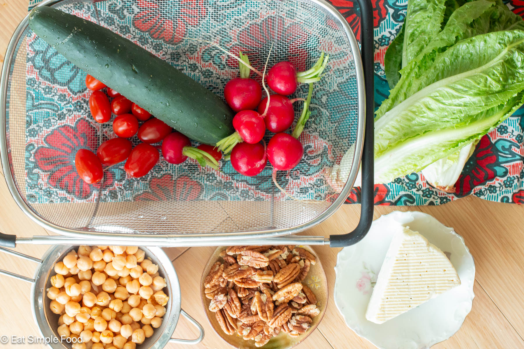 Top down view of a cucumber, radishes, and cherry tomatoes in a strainer. Two heads of romaine lettuce on the side and a strainer of garbanzo beans, small plate of pecans, and blue cheese wedge on the side.