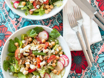 Two shallow white bowls filled with chopped lettuce, sliced cucumbers, sliced radishes, , cherry tomato quarters and blue cheese and pecan crumbles. Two forks, two knives, and two white napkins on the side. Close up.
