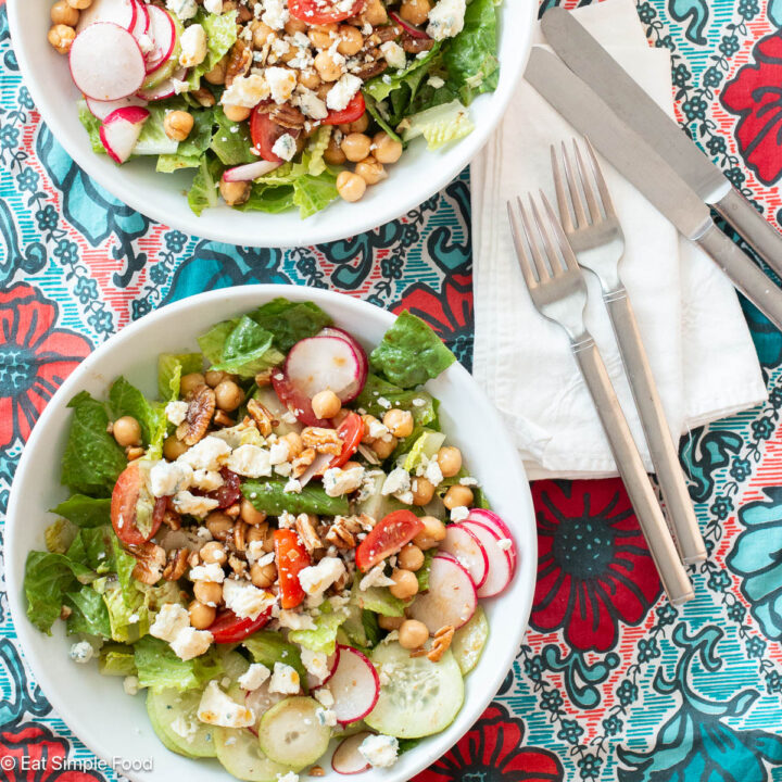 Two shallow white bowls filled with chopped lettuce, sliced cucumbers, sliced radishes, , cherry tomato quarters and blue cheese and pecan crumbles. Two forks, two knives, and two white napkins on the side. Close up.