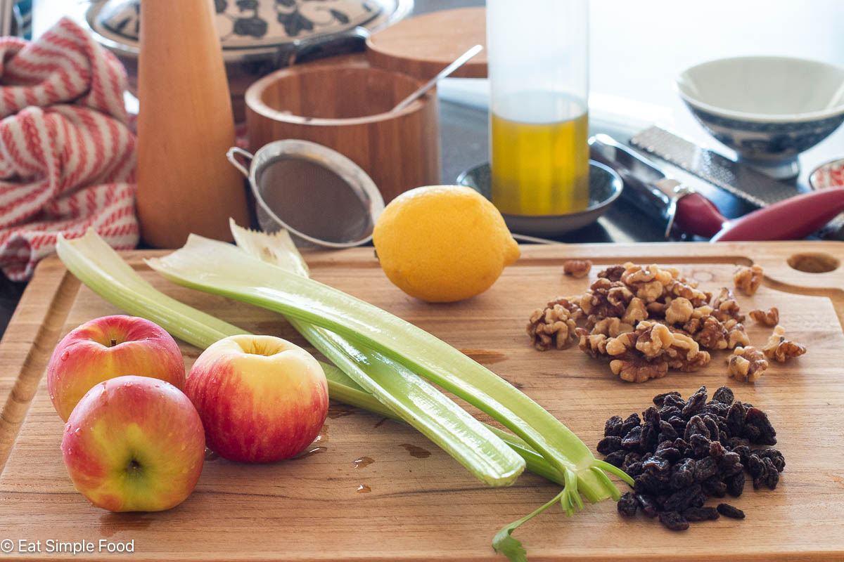 Food items on a wood cutting board: 3 celery stalks, 3 red apples, 1 lemon, mound of walnuts, mound of raisins. Red and white towel in background.