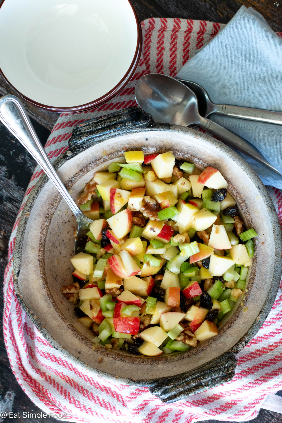 Top view of brown bowl of diced red apples, diced celery, chopped walnuts, and raisins. Spoon sticking out and spoons and white bowls on the side.