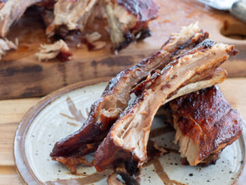 Three pieces of ribs with bone on a white and brown plate with remaining ribs in background on cutting board. Close up,