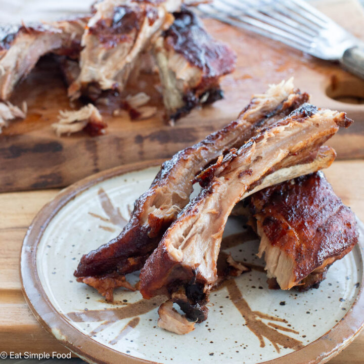 Three pieces of ribs with bone on a white and brown plate with remaining ribs in background on cutting board. Close up,