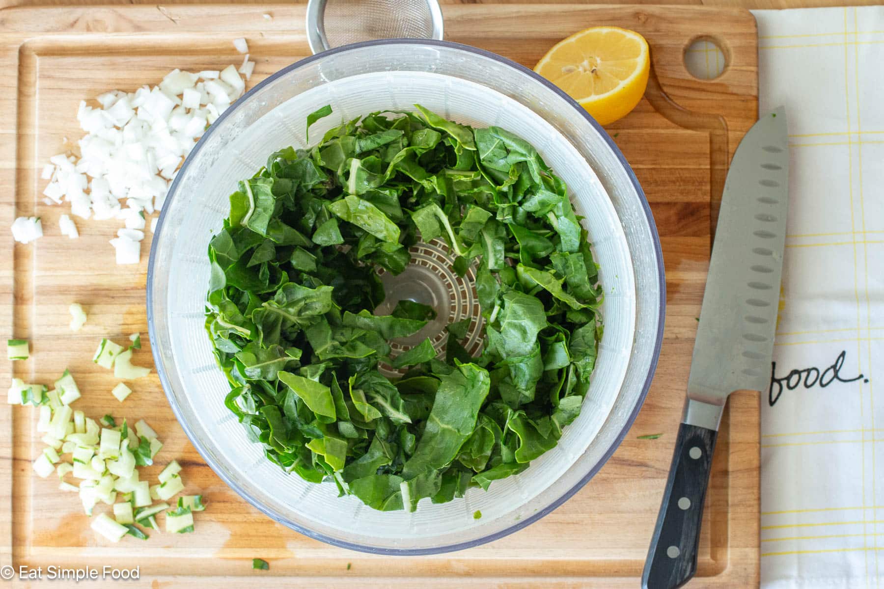 Top down view of a salad spinner filled with chopped greens. Diced onions, diced green stems, ½ a lemon, and a chef's knife on the side.