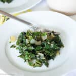 White plate on white tablecloth. Plate has cooked chopped greens and onions and a fork on the side. Close up.