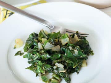 White plate on white tablecloth. Plate has cooked chopped greens and onions and a fork on the side. Close up.