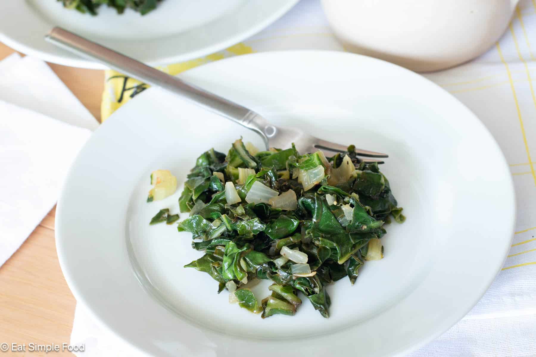 White plate on white tablecloth. Plate has cooked chopped greens and onions and a fork on the side.