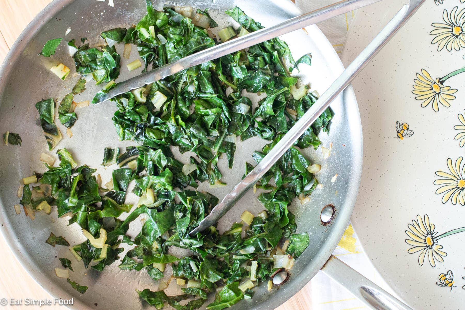 Cooked chopped greens and onions in a stainless steel skillet with metal tongs hanging out. Top down view.