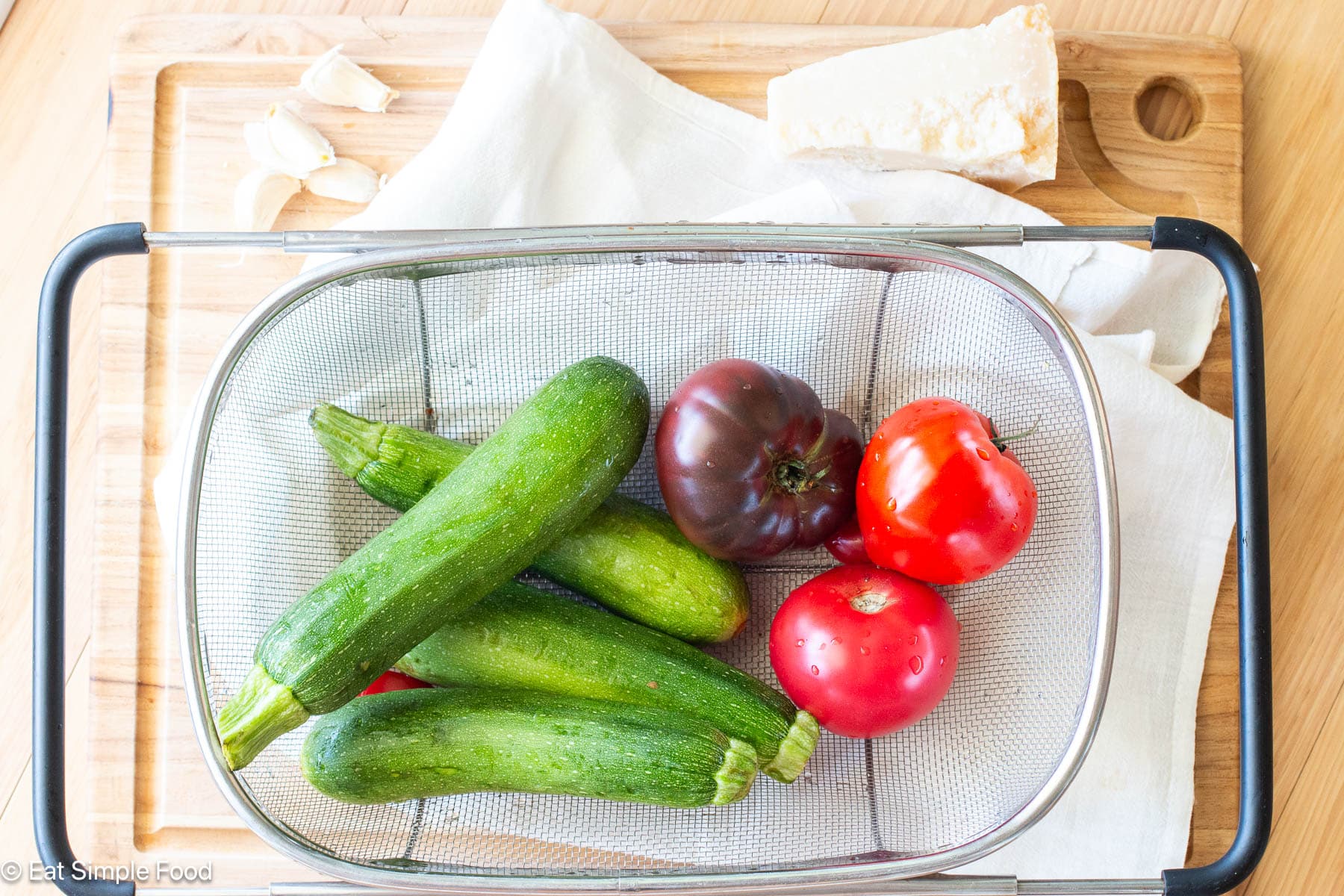 Rectangle colander on white dish towel filled with 4 whole zucchinis and 3 red and purple tomatoes.