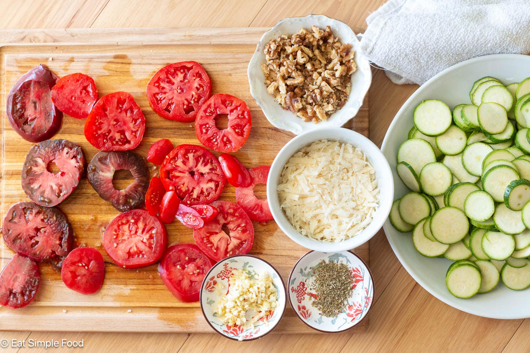 Ingredients on a wood cutting board: sliced tomatoes, grated cheese, chopped nuts, minced garlic, seasoning. Plate of sliced zucchini on side. Top view.