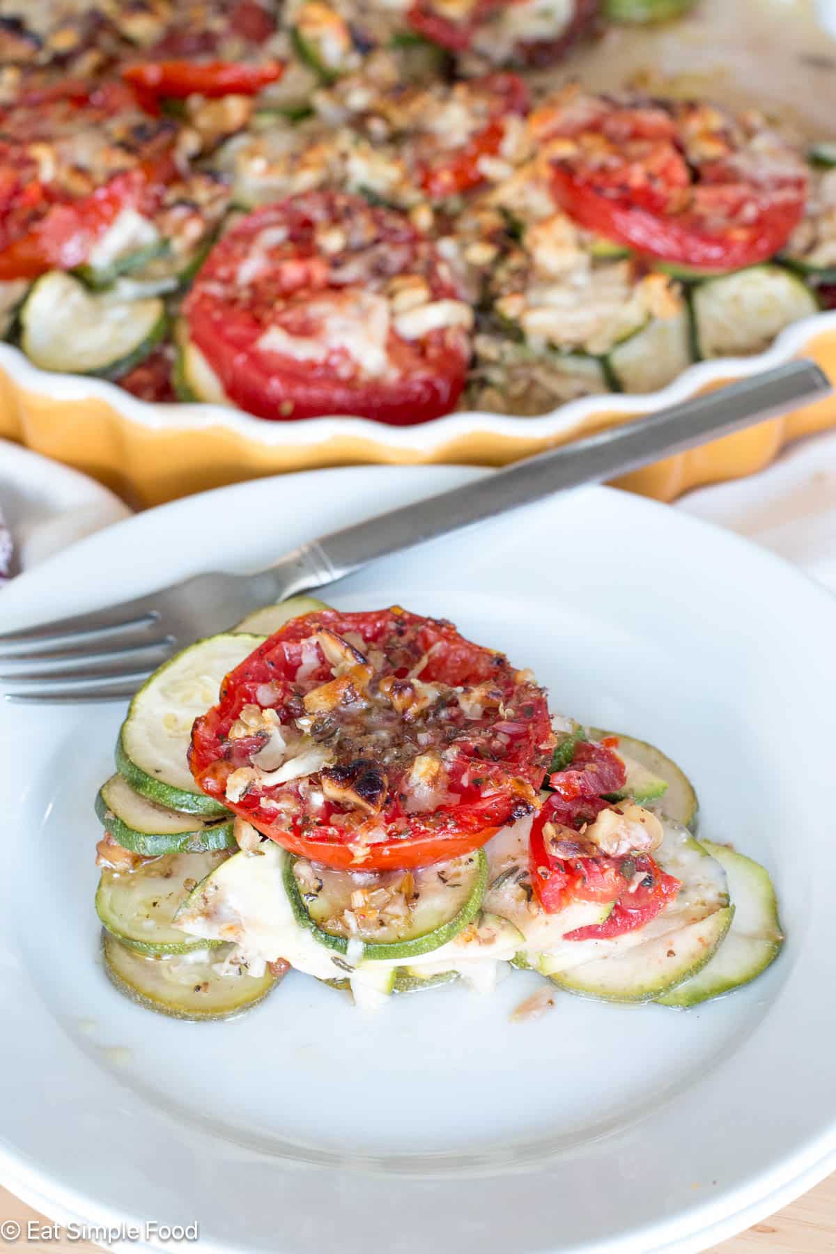 Slice of zucchini and tomato casserole pie on a white plate with knife and large casserole in the background. Side view.