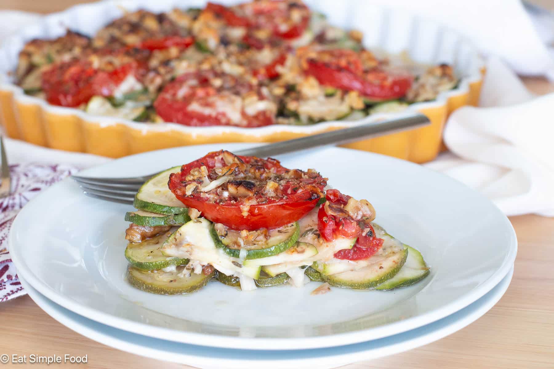 Slice of zucchini and tomato casserole pie on a white plate with knife and large casserole in the background.