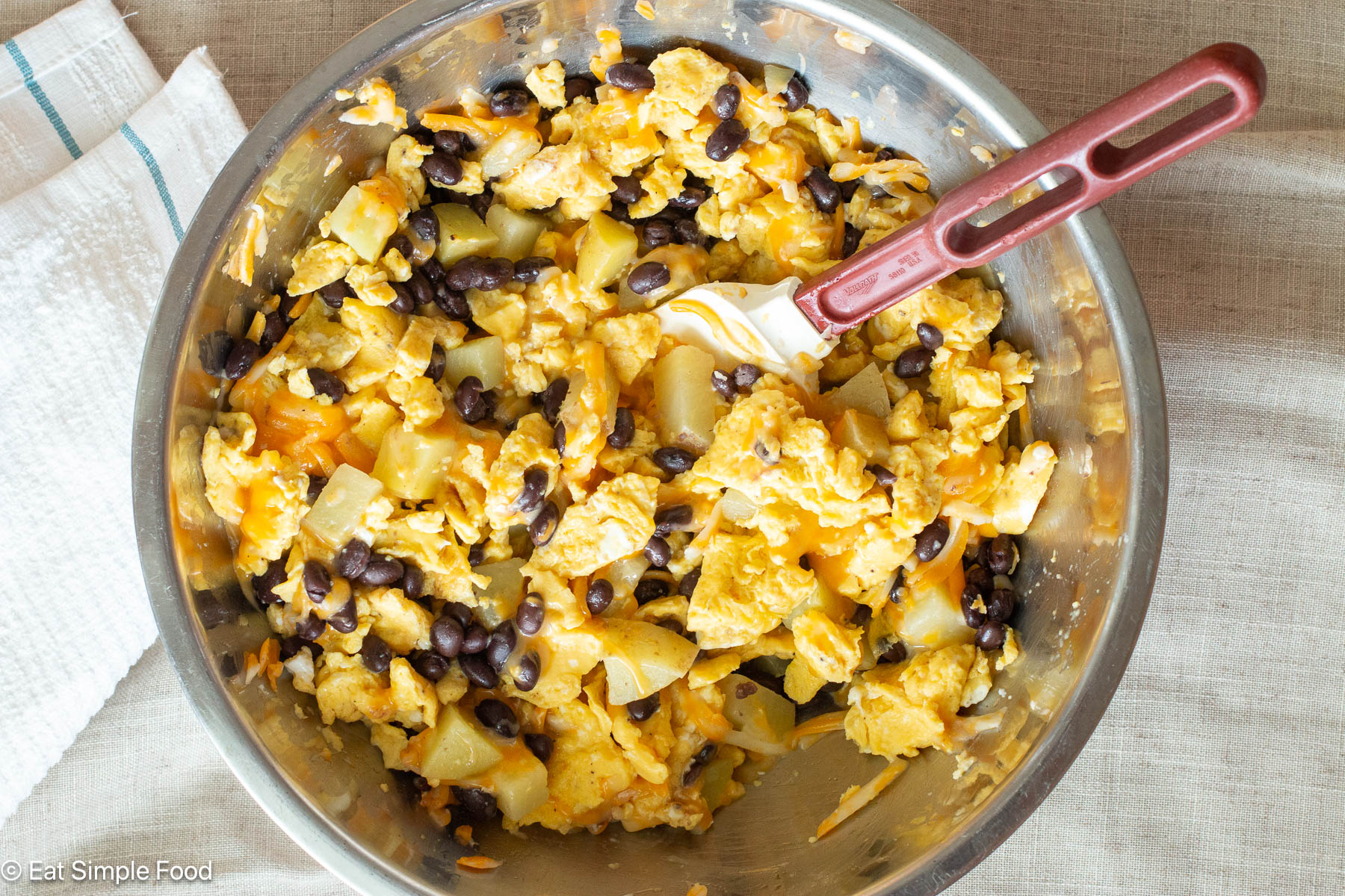 Top down view of stainless steel bowl filled with scrambled eggs, black beans, and potatoes, and a red spatula hanging out the side.