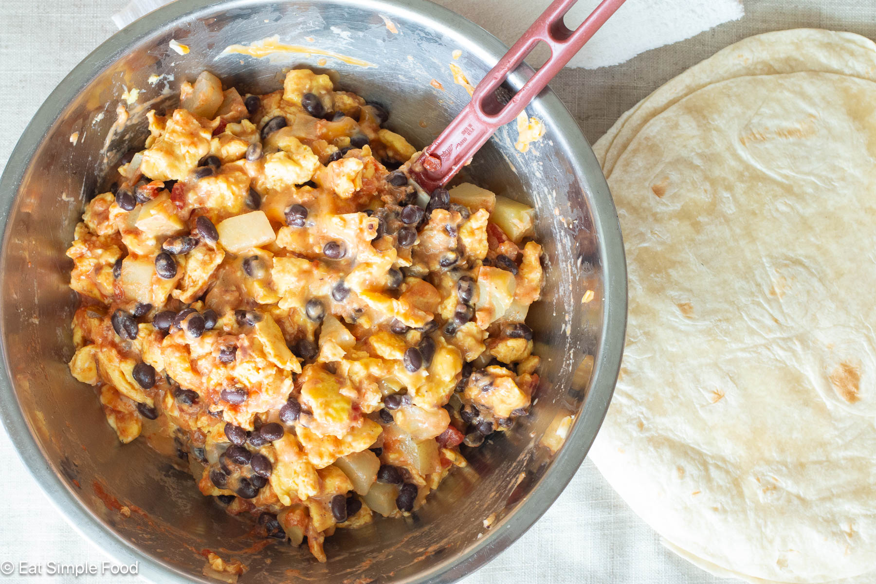Top down view of a stainless steel bowl with scrambled eggs, potatoes, and beans and flour tortillas on the side.