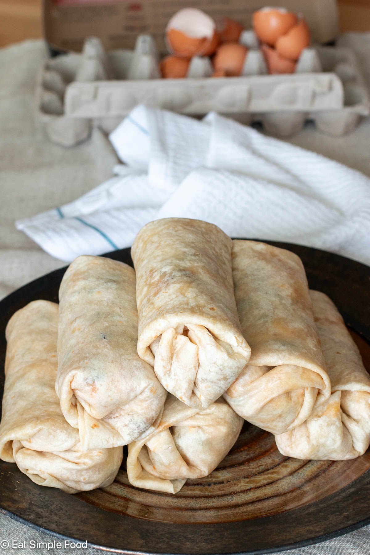 6 stacked burritos on a brown plate with an egg carton with empty eggs in the background.