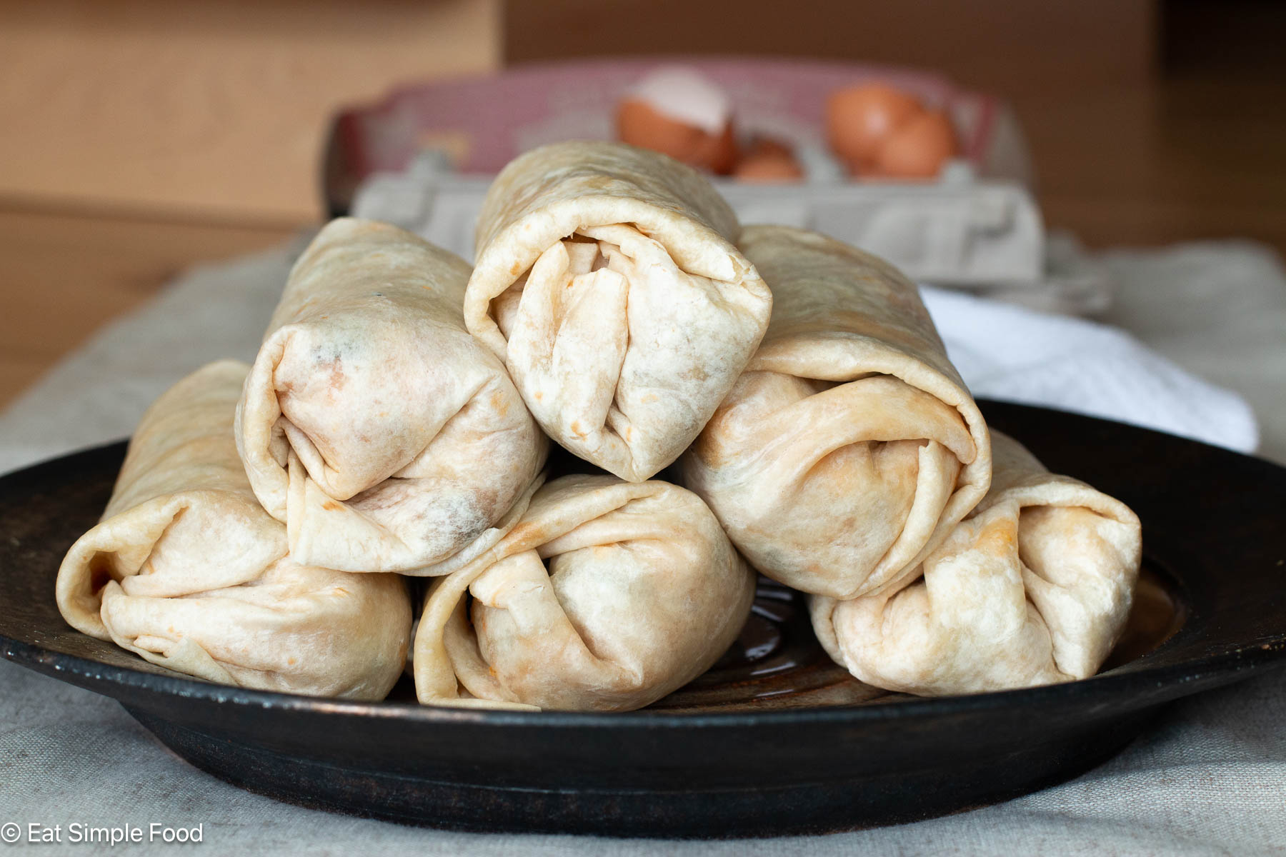 6 stacked burritos on a dark plate with empty egg shells in the background.