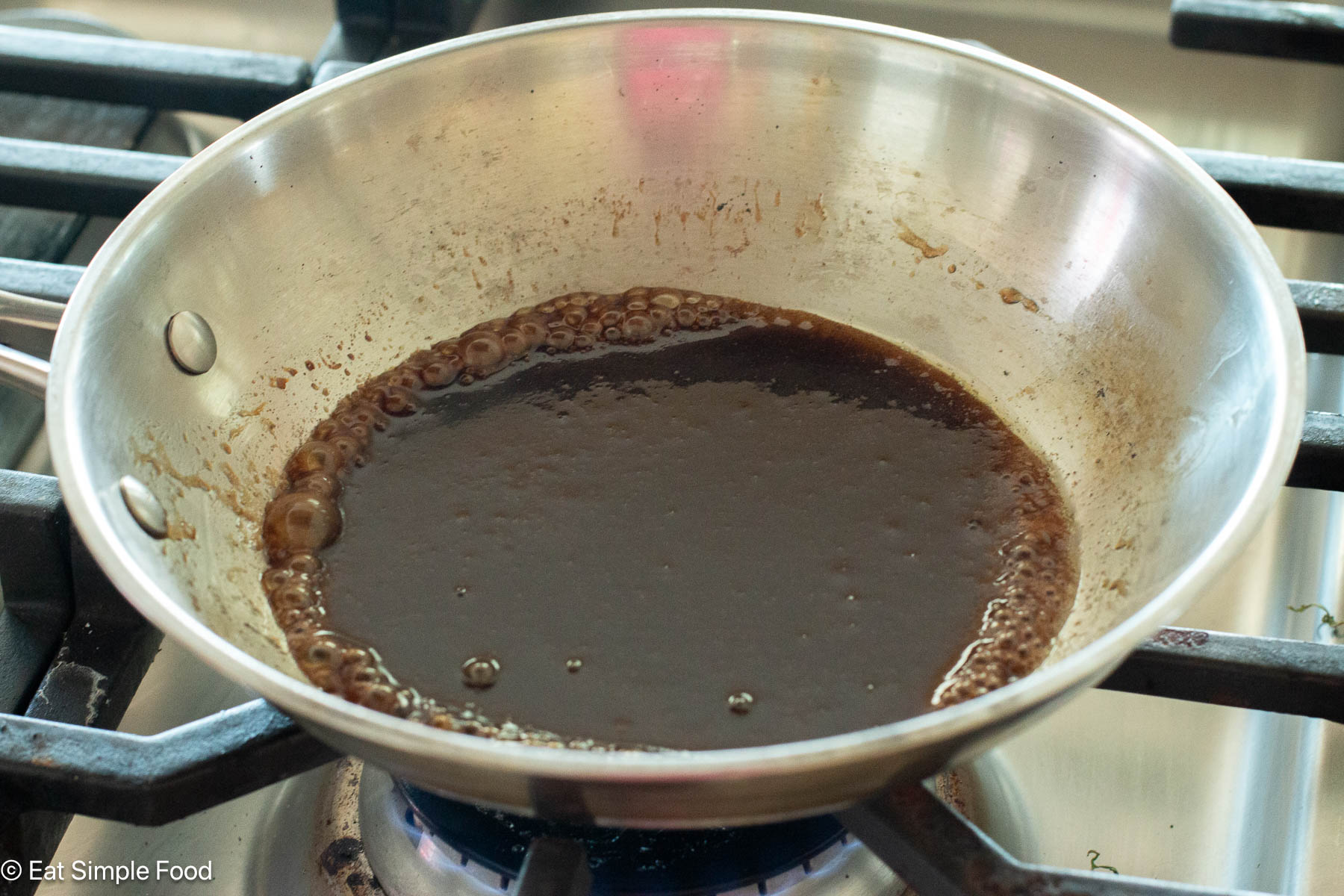 Stainless steel pan of brown sauce simmering on a stovetop gas range.