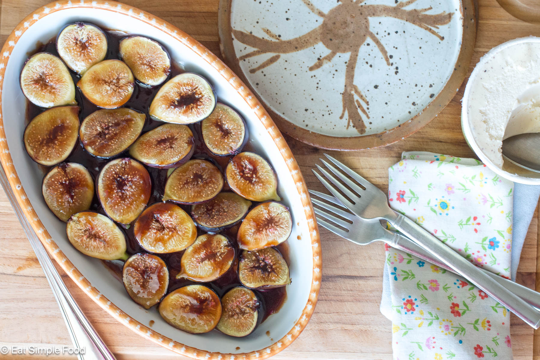 Top down view of fig halves in oval baking dish with brown sauce. Two fork, napkins, and plates on the side.