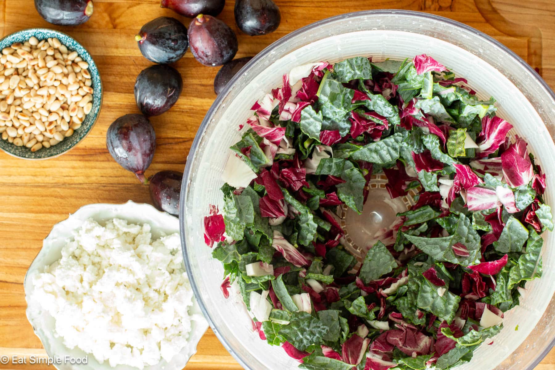 Top down view of chopped up radicchio and kale in a salad spinner on a wood cutting board with whole figs, crumbled cheese, and nuts on the side.