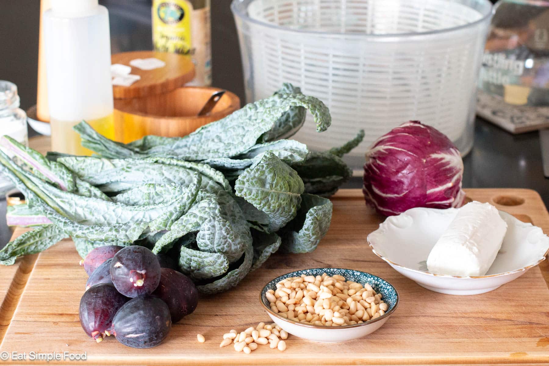 Ingredients on a wood cutting board: kale leaves, radicchio, log of goat cheese, pine nuts ina small bowl, and whole figs. Salad spinner in background.