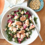 Top down view of a white plate with chopped greens with quartered figs and pine nuts and goat cheese. Fork on the side of plant and pine nuts on the side.