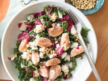Top down view of a white plate with chopped greens with quartered figs and pine nuts and goat cheese. Fork on the side of plant and pine nuts on the side.