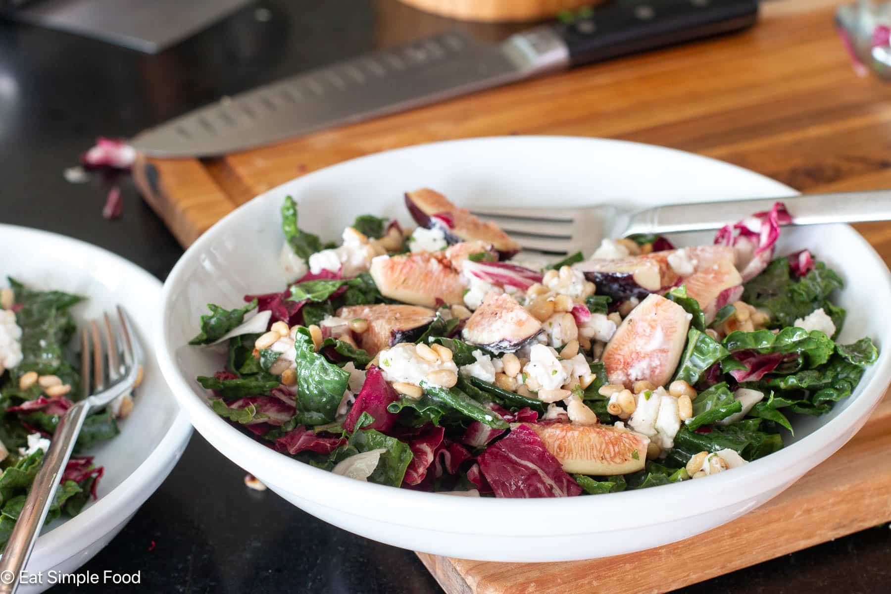 One white plate with chopped greens with quartered figs and pine nuts and goat cheese crumbles on a wood cutting board. Fork hanging out the side of plate.