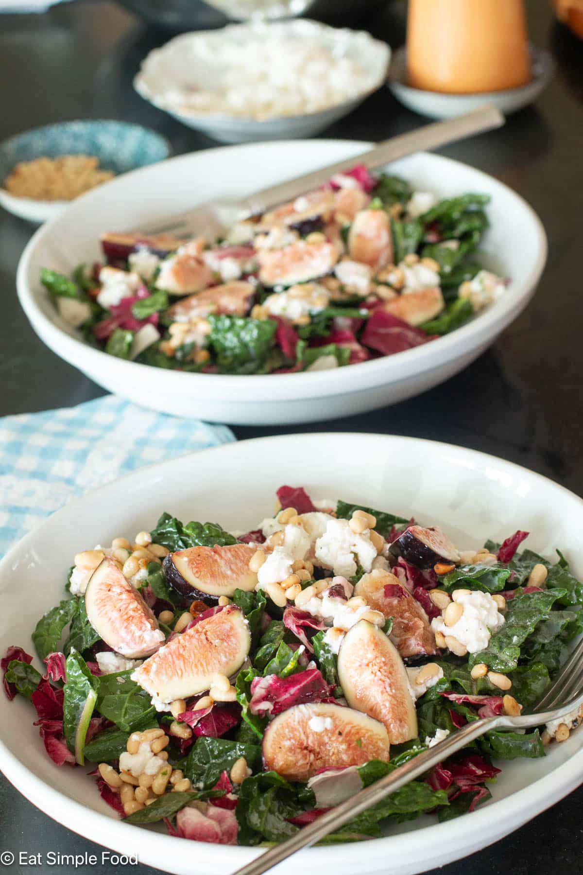 Two white plates with chopped greens with quartered figs and pine nuts and goat cheese. 2 Forks hanging out the side.