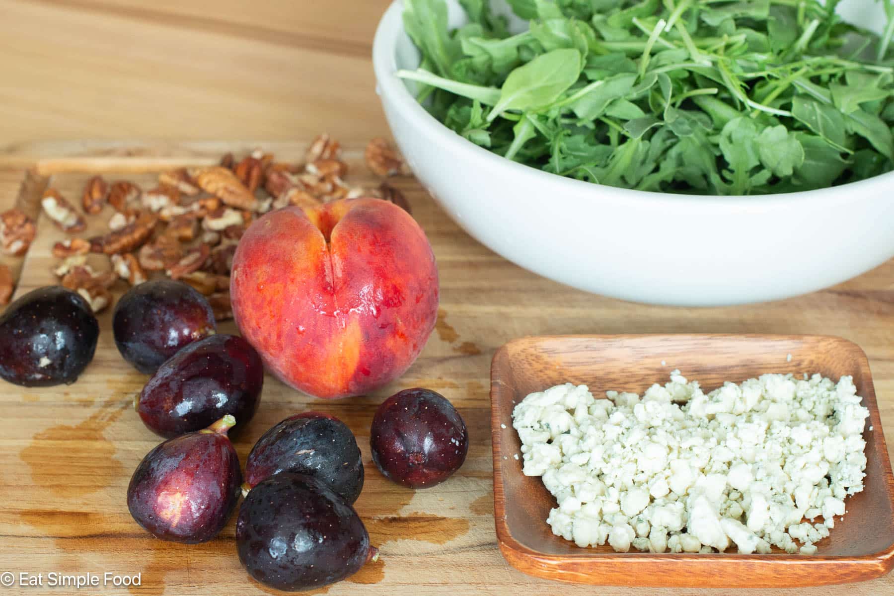 Ingredients on wood cutting board: seven whole figs, 1 peach, pecan pieces, crumbled blue cheese, and a white bowl filled with arugula.