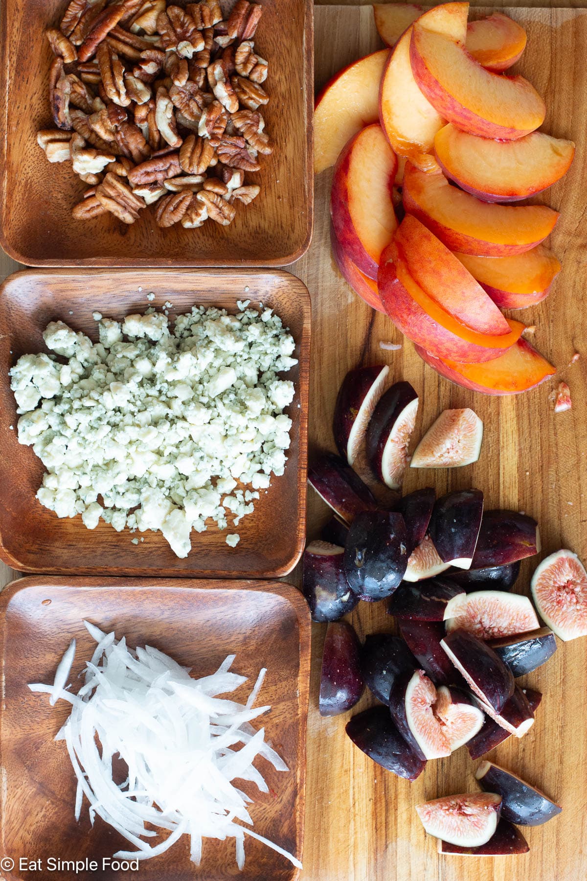Top down view of ingredients on a wood cutting board: pecan pieces, crumbled blue cheese, sliced white onions, quartered pink figs, peach slices.