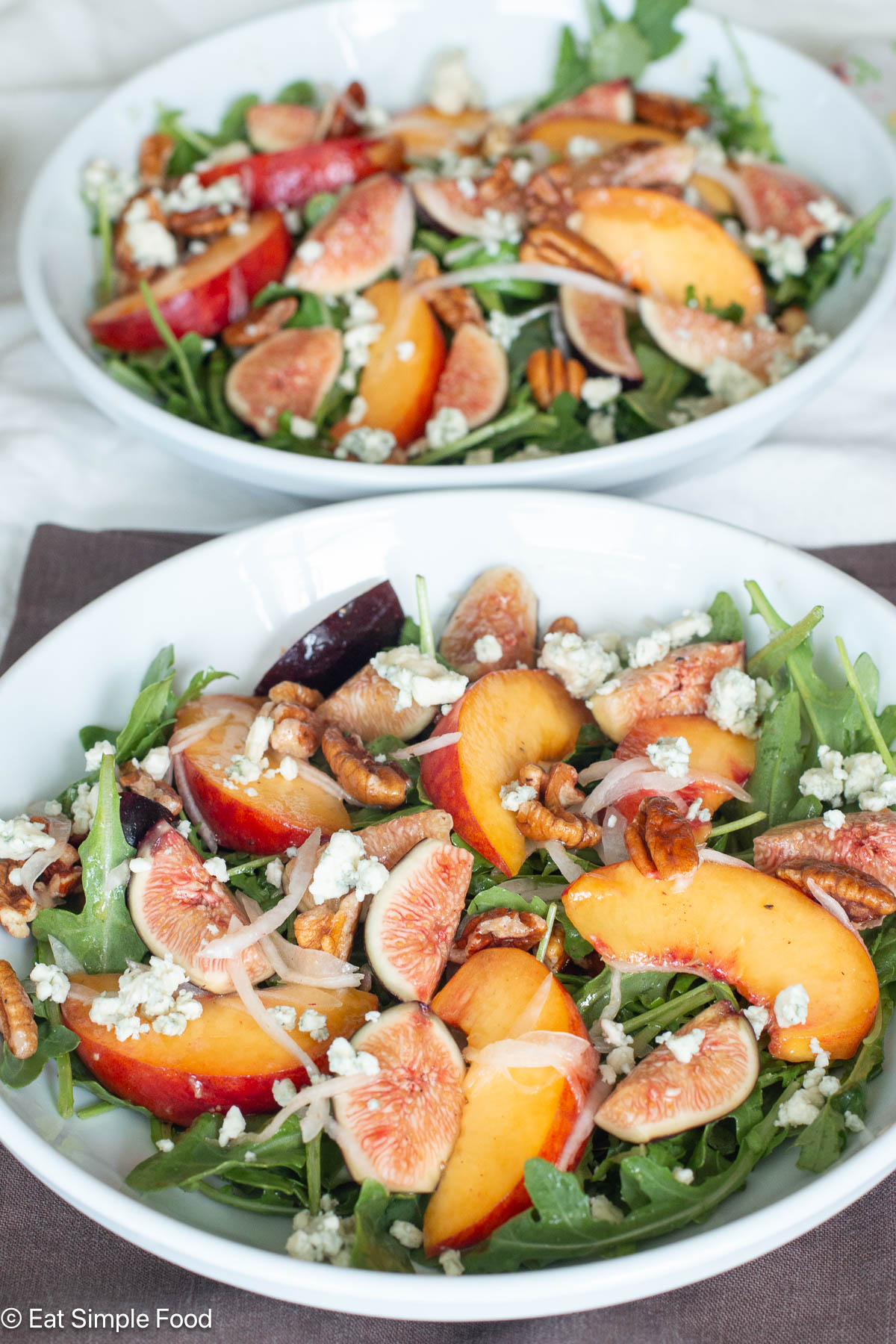 Two white plates with green salad, sliced peaches, quartered figs, white cheese crumbles and sliced onions.