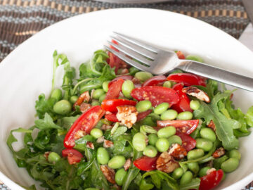 Side view of white bowl of green salad with shelled edamame, pecans, cherry tomato halves. Fork sticking out.