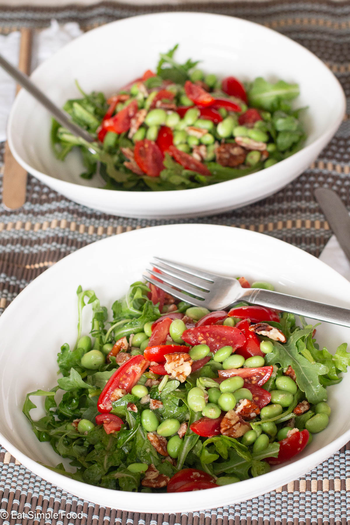 Side view of two white bowls of green salad with shelled edamame, pecans, cherry tomato halves. Forks sticking out.