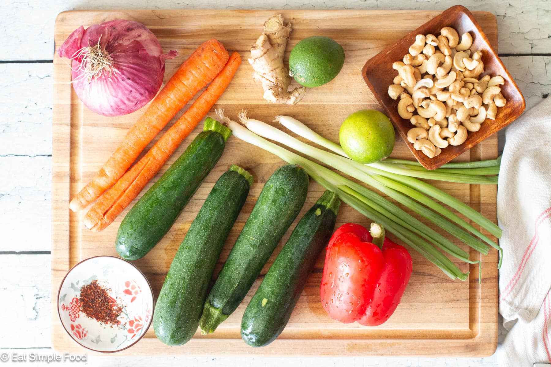 Ingredients on a wood cutting board. Top down view. 4 zucchini, 2 carrots, 1 red onion, knob of ginger, 2 limes, container of cashews, container of red pepper chili flakes.