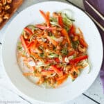 Top view of zucchini noodles with carrots, red onions, red peppers, green onions and cashews on a white plate with chopsticks.