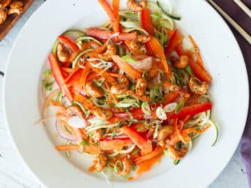Top view of zucchini noodles with carrots, red onions, red peppers, green onions and cashews on a white plate with chopsticks.