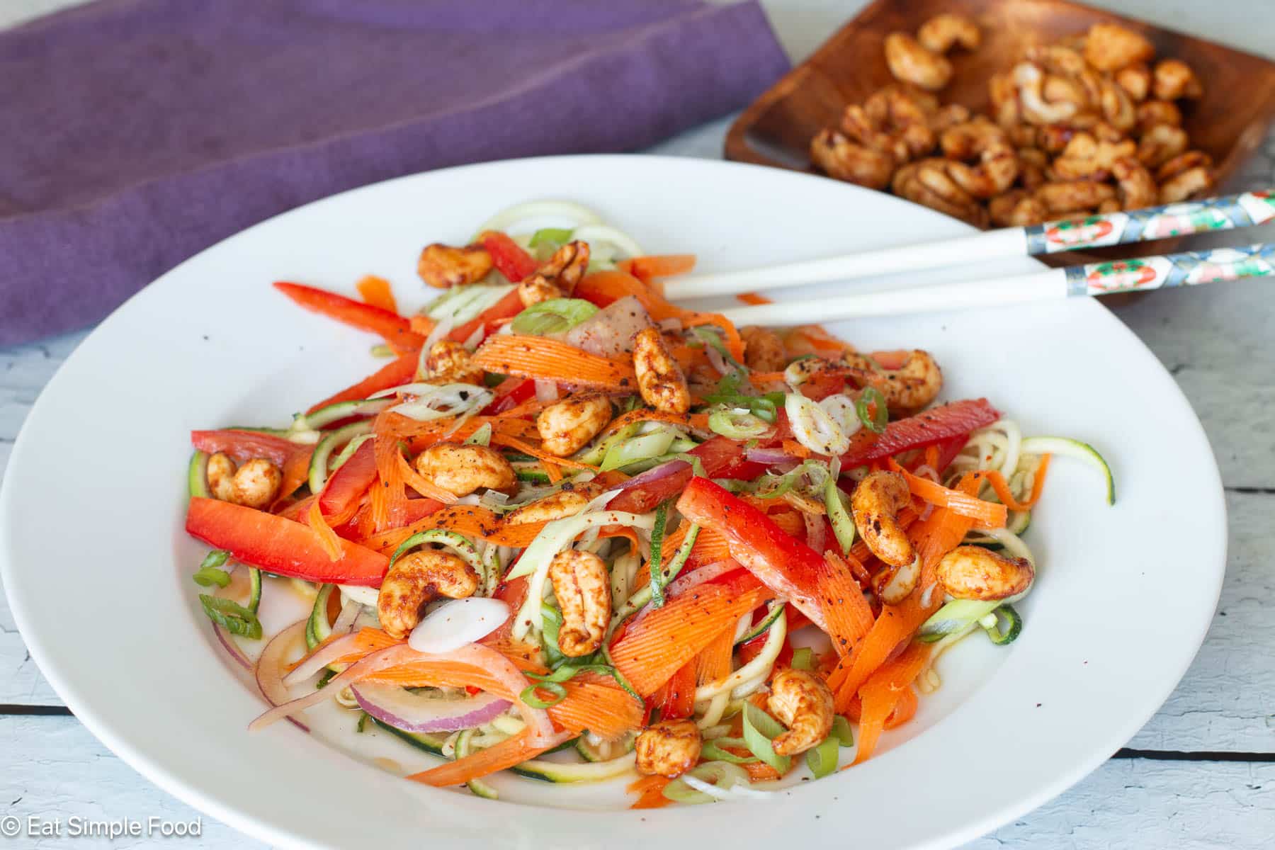 Side view of zucchini noodles with carrots, red onions, red peppers, green onions and cashews on a white plate with chopsticks.