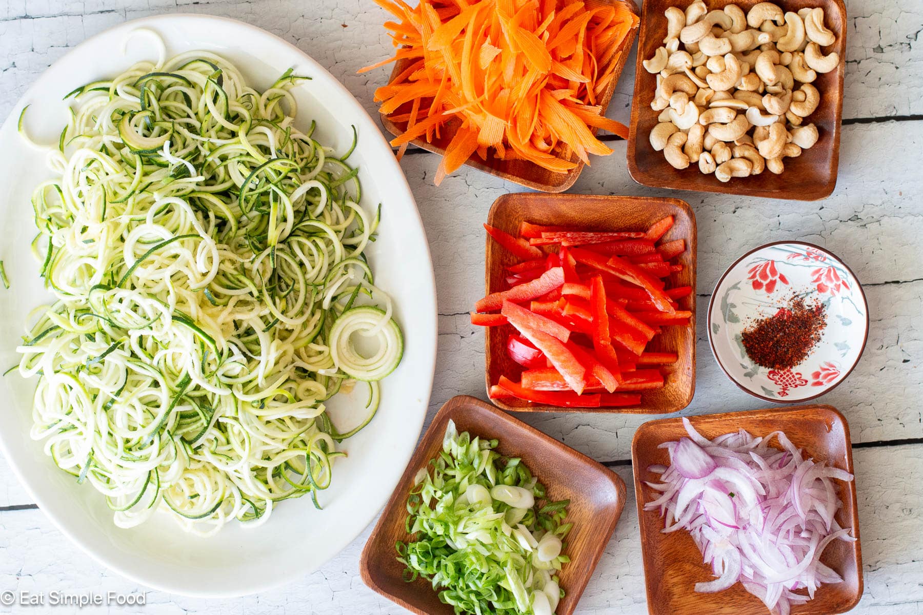 Top down view of a white plate of zucchini noodles and 5 wood bowls with different ingredients: shaved carrots, sliced red peppers, cashews, sliced green onions, sliced red onions.