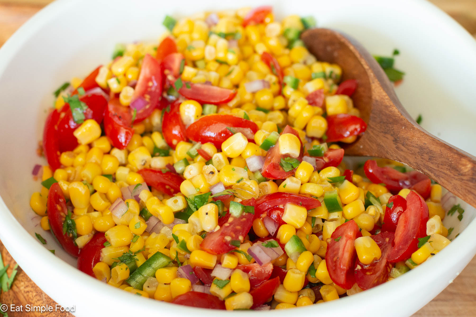 White bowl of mixed corn, diced tomatoes, diced jalapeno, minced cilantro with a wood spoon hanging out of bowl.