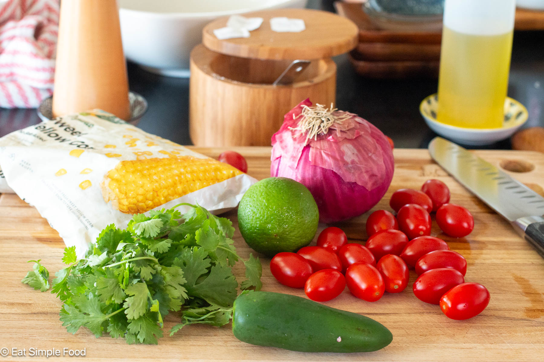Ingredients on wood cutting board: jalapeno, lime, cherry tomatoes, bunch of cilantro, red onion, bag of frozen corn.